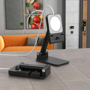 A foldable phone stand in black with an attached light, positioned on a table with fruit in the background.