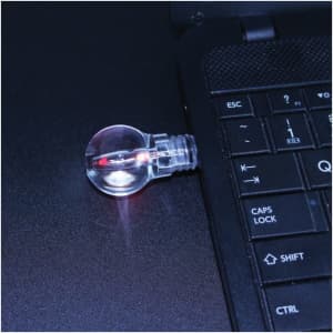 A light bulb flash drive in a clear casing connecting to a laptop, glowing with a red light.