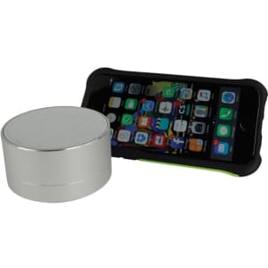 A silver Bluetooth speaker next to a smartphone with a protective case, displaying various app icons.