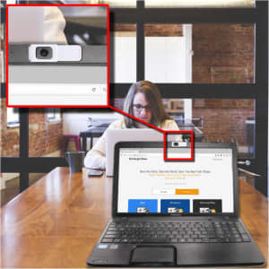 A webcam cover slide in white positioned on a laptop, with a blurred background of a woman working at a desk.