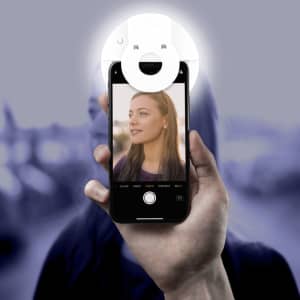 A hand holds a smartphone with a circular white Smiley LED Selfie Light attached, illuminating a person's face in the photo.