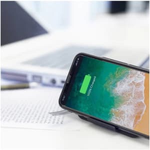 A wireless charger stand with a black finish, holding a smartphone displaying a charging icon, beside a laptop and documents.