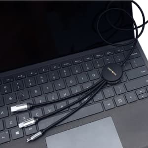 A black charging cable with multiple connectors lies on a laptop keyboard. The cable features a circular connector with branding.