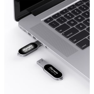 A silver Olympia LED flash drive is plugged into a laptop's USB port, with another flash drive beside it.