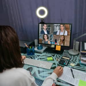 A workspace with a person in front of a computer displaying a video call, featuring a ring light and various office supplies.