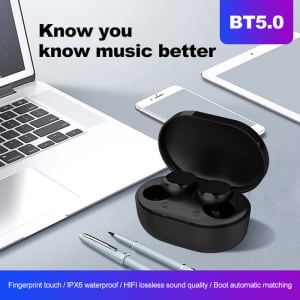 A pair of black wireless earbuds in a charging case, positioned beside a laptop and smartphone on a desk.