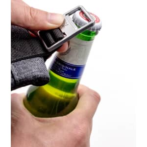 A hand holding a bottle with a grey backpack strap, using a bottle opener attached to the strap.
