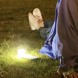 A smartphone torch emits a warm light on the grass, with a person in casual attire sitting nearby.
