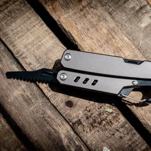 A multi-tool with a silver body, featuring a small saw blade, resting on a wooden surface. It includes a keyring.
