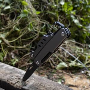 A black multi-tool featuring various tools, including a knife, stably positioned on a wooden surface amidst foliage.