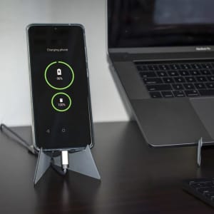 A transparent phone stand displaying a charging smartphone beside a laptop on a desk.