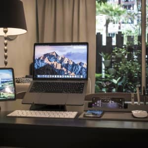 A sleek grey Ascend Mini Stand displays a laptop and phones, set on a stylish desk with greenery in the background.