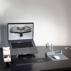 A sleek grey Ascend Mini Stand holding a smartphone, positioned beside a laptop, keyboard, and pen holder on a desk.