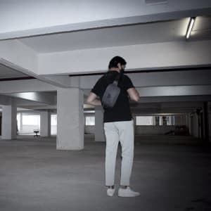 A man in a black shirt and light trousers is carrying a grey anti-theft sling bag in a parking garage.