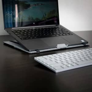 A grey laptop stand supports a laptop on a dark wooden desk, with a white keyboard nearby.