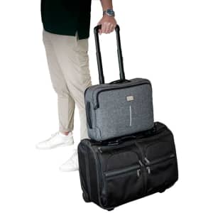 A grey laptop bag with wheels is stacked on a black wheeled suitcase, held by a person in casual attire.