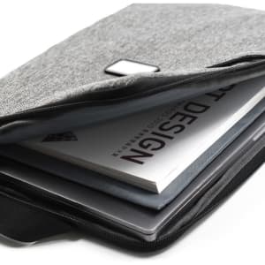 A grey laptop sling bag with a zip, containing a laptop and a design book. It features a small logo.