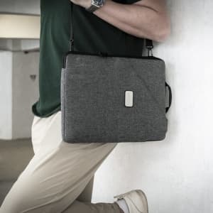 A grey laptop sling bag with a handle and adjustable strap worn by a person. The bag features a logo.