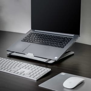A grey laptop stand with a laptop, white keyboard, and mouse on a dark wooden desk.