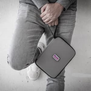 A grey travel wallet with a strap, featuring a logo, held against a person's leg dressed in denim.