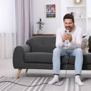 A man seated on a grey couch using a phone, with a charging cable visible on the floor. The setting is bright and modern.