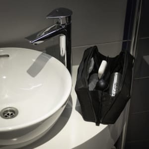 A black toiletry bag with various grooming items beside a white sink and chrome tap. The bag features a logo.