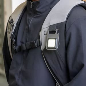 A compact black multi-tool attached to a backpack strap, featuring a bright LED light.