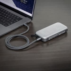 A silver Bluetooth speaker power bank with a textured surface, connected to a laptop with a braided cable.