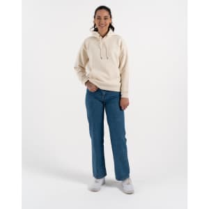 A woman in a beige hoodie and blue jeans stands on a light background, wearing white sneakers. The hoodie has a logo.