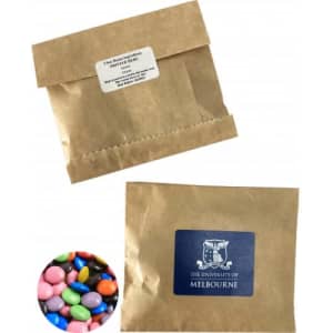 A Kraft paper bag containing colourful choc beans, with a logo on the front. The bag is brown and sealed at the top.
