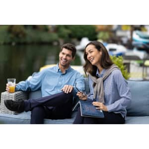 A couple sits outdoors, both wearing light blue gingham shirts, enjoying drinks, with greenery and water in the background.