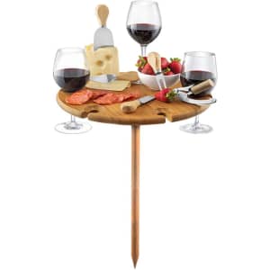 A wooden outdoor party table with a stake, holding wine glasses, cheese, strawberries, and utensils.