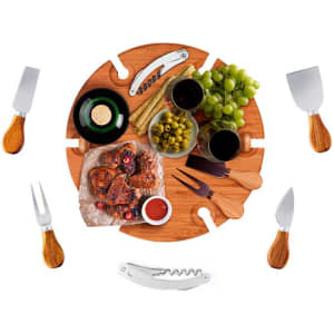 A wooden circular serving board features cheese, fruits, wine, and utensils, surrounded by knives and a corkscrew.