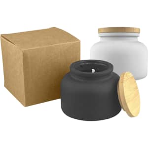 A black candle jar with a wooden lid next to a white candle jar, both with a brown box.