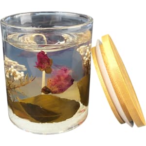 A floral candle in a clear glass jar features dried pink roses and white flowers, with a wooden lid nearby.
