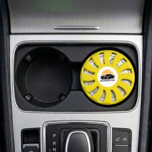 A round, yellow air freshener is placed in a car cup holder, featuring a logo and a vented design.