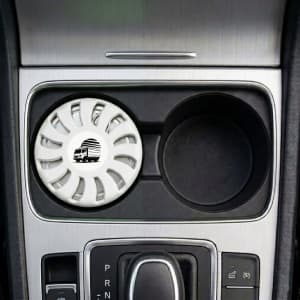 A white air freshener sits in a car cup holder, featuring a circular design and a logo.