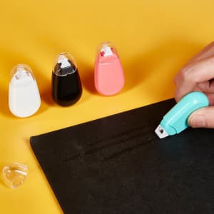 A hand using a turquoise Bayla Glue Tape on a black surface, with white, black, and pink glue tape holders in the background.