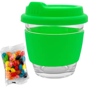A reusable coffee cup with a clear glass body and a bright green silicone lid and sleeve, alongside a packet of colourful jelly beans.