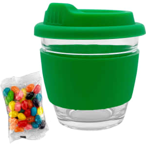 A reusable glass coffee cup with a green silicone sleeve and lid, accompanied by a bag of colourful jelly beans.