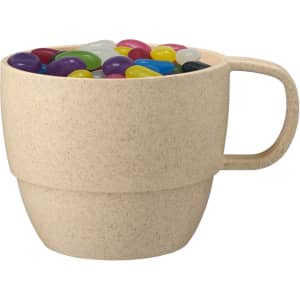 A reusable wheat straw cup in a light beige colour filled with colourful jelly beans. It features a handle and a logo.