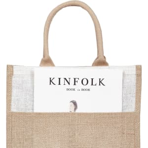 A jute bag in natural brown with handles, featuring a magazine partially visible in the front pocket and a logo.