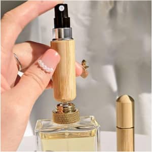 A mini bamboo atomiser held in a hand, with a perfume bottle in the background. The product features a natural bamboo finish.