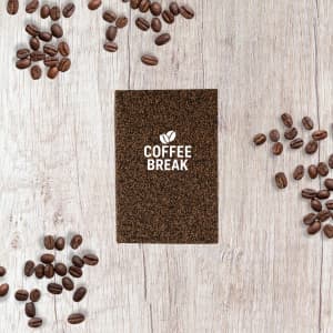 A brown A6 notebook surrounded by coffee beans on a wooden surface, featuring a logo on the cover.