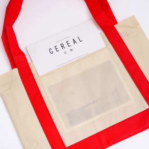 A beige reusable grocery bag with red handles features a clear pocket, holding a magazine inside.