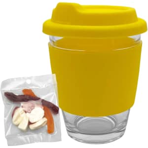 A reusable glass coffee cup with a yellow silicone lid and sleeve, accompanied by a clear bag of assorted jelly sweets.