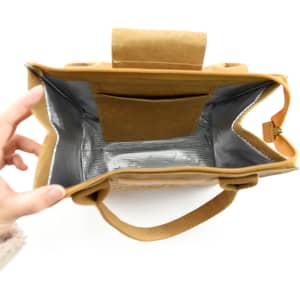 A tan lunch bag with a silver interior and a small pocket, held open by a hand. The bag has padded handles.