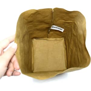 A large brown kraft paper storage bag with a squared base and a logo inside. The bag is held by a hand.