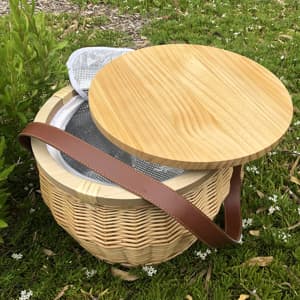 A round wicker picnic cooler basket in natural tones, featuring a wooden lid and a brown leather strap.