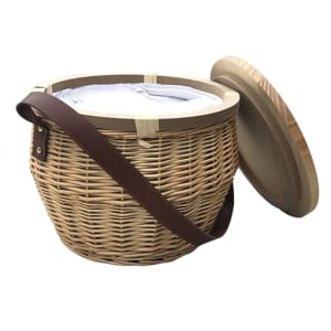 A round wicker picnic cooler basket in natural tones with a wooden lid and a brown strap. It features a logo.